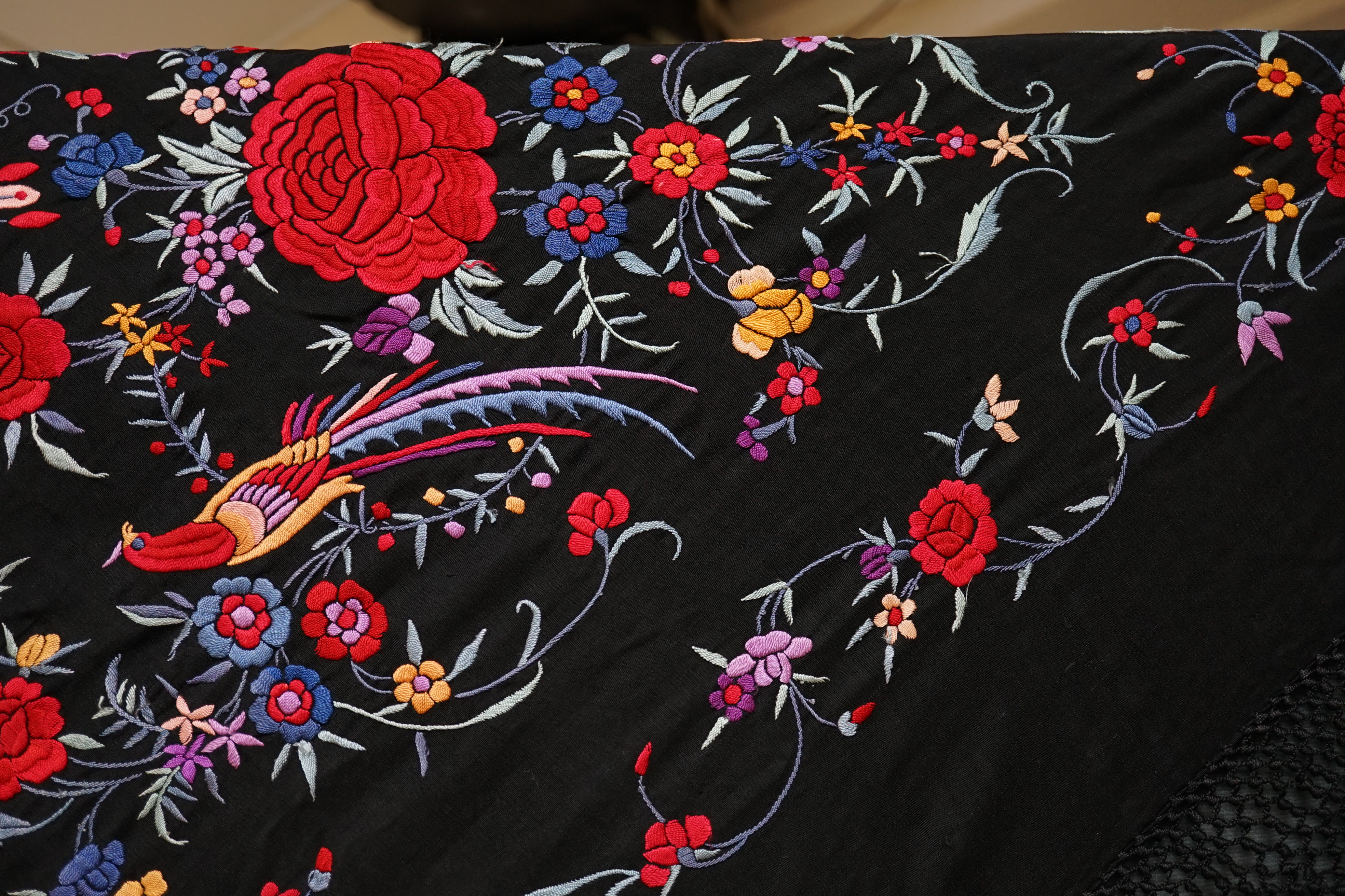 An early 20th century Chinese black silk fringed shawl with polychrome silk embroidery, embroidered with meandering floral design, the corners embroidered with three brightly coloured birds of paradise, large red roses a